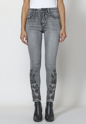 Slim-fit grå jeans med sort blomsterbroderi på underbenene, kombineret med sorte ankelstøvler og en hvid top, båret af en stående person.