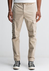 Man in een beige cargobroek en zwarte hoge sneakers, staande tegen een effen lichte achtergrond.