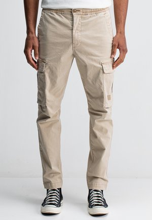 Mann in beigen Cargo-Hosen und schwarzen High-Top-Sneakern, der vor einem schlichten hellen Hintergrund steht.