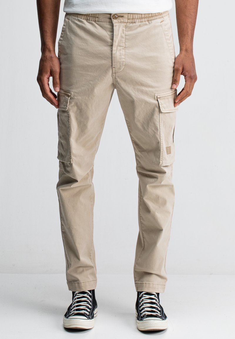 Man in een beige cargobroek en zwarte hoge sneakers, staande tegen een effen lichte achtergrond.