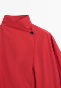 High collar red garment with a single black button near the neckline and overlapping fabric design.
