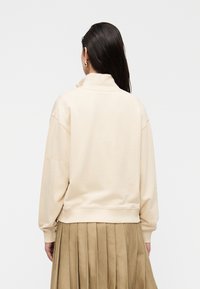 Κρεμ φούτερ με ψηλό γιακά, χαλαρή εφαρμογή στους ώμους και ριμπ τελείωμα, σε συνδυασμό με φούστα μπεζ με πιέτες.