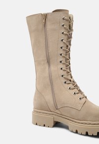 Beige suede combat boot with a lace-up front, side zipper, chunky sole, and contrasting stitching detail. Textured material with a rugged design.