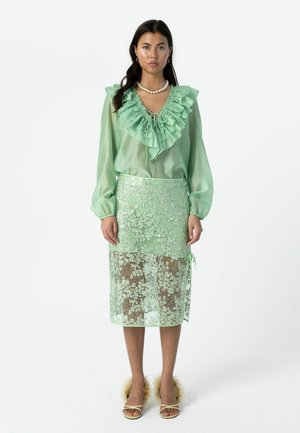 Nő áll, világos zöld áttetsző fodros blúzt, hozzá illő virágos csipkerockot, gyöngysort, karika fülbevalót, valamint nyitott orrú, szőrmés részletekkel díszített magas sarkút viselve.