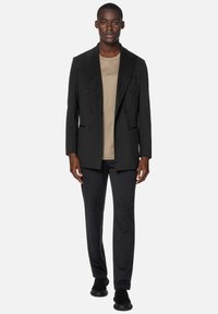 Čierny dvojradový blazer s vykrojenými klopami, skombinovaný s béžovým tričkom s okrúhlym výstrihom a tmavými nohavicami. Model má na sebe elegantné čierne topánky.