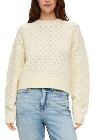 Cremefarbener gestrickter Pullover mit Rautenmuster, runder Ausschnitt, überschnittenen Schultern und geripptem Saum. Kombiniert mit hellblauen Jeans.