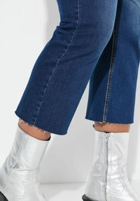 Tummansiniset farkut, joissa on hapsut ja suora leikkaus, yhdistettynä hopeisiin metallisiin nilkkureihin, joissa on sivuvetoketju.