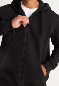 Zwarte zip-up hoodie van zacht stof, met kangoeroe pockets, geribbelde manchetten en een verstelbare trekkoordcapuchon.