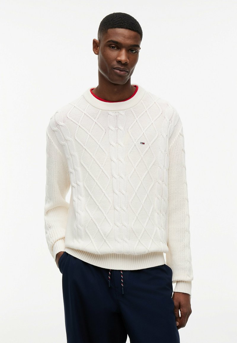 Jeune homme portant un pull en maille torsadée blanc avec un petit logo et un pantalon bleu marine, debout avec une main dans sa poche.