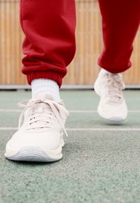 Lyse beige sportsko med tekstureret mesh-overdel og flade snøringer, båret med hvide sokker og røde sweatpants, på en grøn overflade.