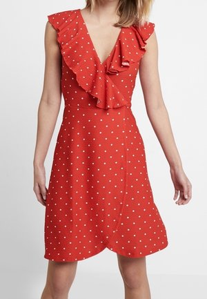 Femme portant une robe rouge sans manches à pois blancs avec un col en V volanté, debout devant un fond clair uni.