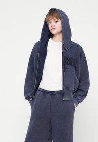 Mladý človek v tmavomodrej oversize mikine na zips s kapucňou, bielej košeli a zodpovedajúcich tmavomodrých nohaviciach, s rukami v vreckách.