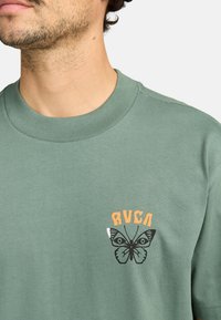 Camiseta de algodón verde con cuello redondo acanalado, con un gráfico de mariposa y el texto "RVCA" en naranja encima del diseño, en el área del pecho izquierdo.
