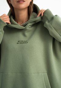 Przesadnie duża zielona bluza z kapturem, wykonana z miękkiego materiału, z dużą przednią kieszenią oraz wyszytym napisem "BĄDŹ UNIKALNY, BĄDŹ SOBĄ" na klatce piersiowej.