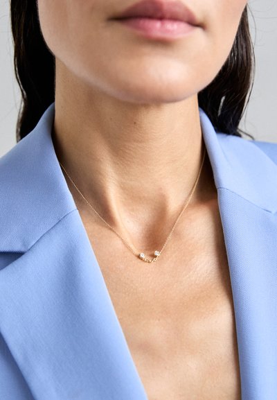 Collar de oro con dos acentos circulares claros, llevado sobre un blazer azul claro. La vista en primer plano resalta el delicado diseño y textura de la cadena.