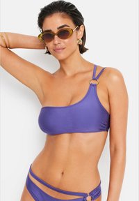 Violet bikini viršus su viena petneša, puošniu auksiniu žiedu ir švelnia, tampri audiniu. Modelis derina jį su akinių nuo saulės.