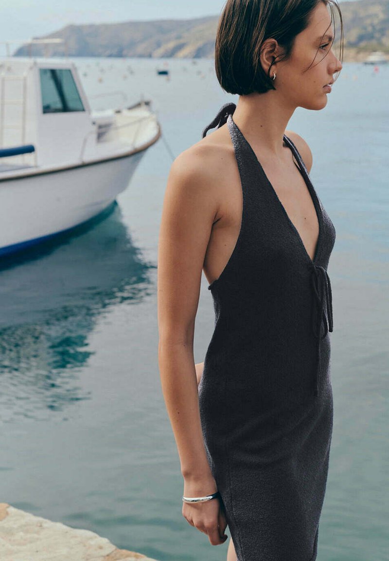 Femme en robe noire sans manches debout près d'un quai avec un bateau blanc et une eau calme en arrière-plan.
