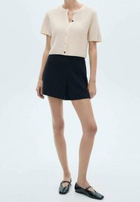 Kremowy kardigan z krótkim rękawem o fakturze jednolitej, zapinany na guziki, w połączeniu z ciemnonavym spodniami szortami w wersji dopasowanej, noszony z czarnymi płaskimi butami.