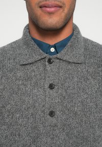 Γκρι πλεκτό πουλόβερ με γιακά, με τέσσερα μαύρα κουμπιά και μπλε πουκάμισο με κουμπιά να φαίνεται εν μέρει από κάτω.