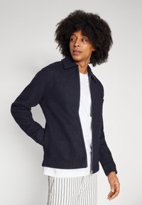 Navy wool blend jacket featuring a zip front, collared neckline, and a single chest pocket, paired with a white crew neck shirt.