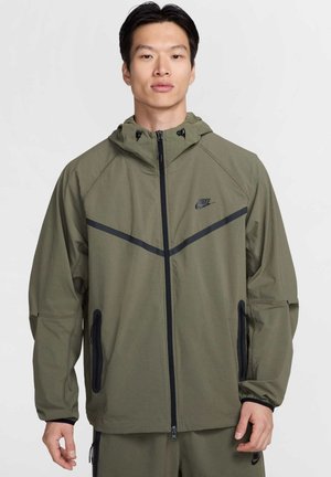 Chaqueta Nike en verde olivo con cierre de cremallera, capucha y detalles en negro. Presenta un diseño geométrico y bolsillos laterales con cremalleras.