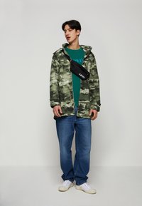 Zöld álcázó mintás dzseki kapucnival, kék pólóra rétegezve. Kék farmernadrággal és fehér sportcipővel viselve. Fekete keresztpántos táska.