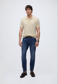 Homme debout portant un polo beige à manches courtes, un jean bleu avec une ceinture marron, des mocassins en cuir marron à pampilles, et une montre-bracelet.