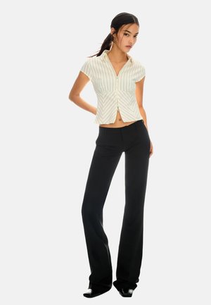 Witte blouse met dunne groene strepen, kraag en rits. Zwarte flared broek met een nauwsluitende pasvorm. Model staat in een zelfverzekerde pose.