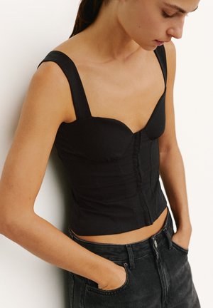 Femme portant un haut noir moulant sans manches avec des boutons sur le devant et un jean taille haute foncé, les mains dans les poches.