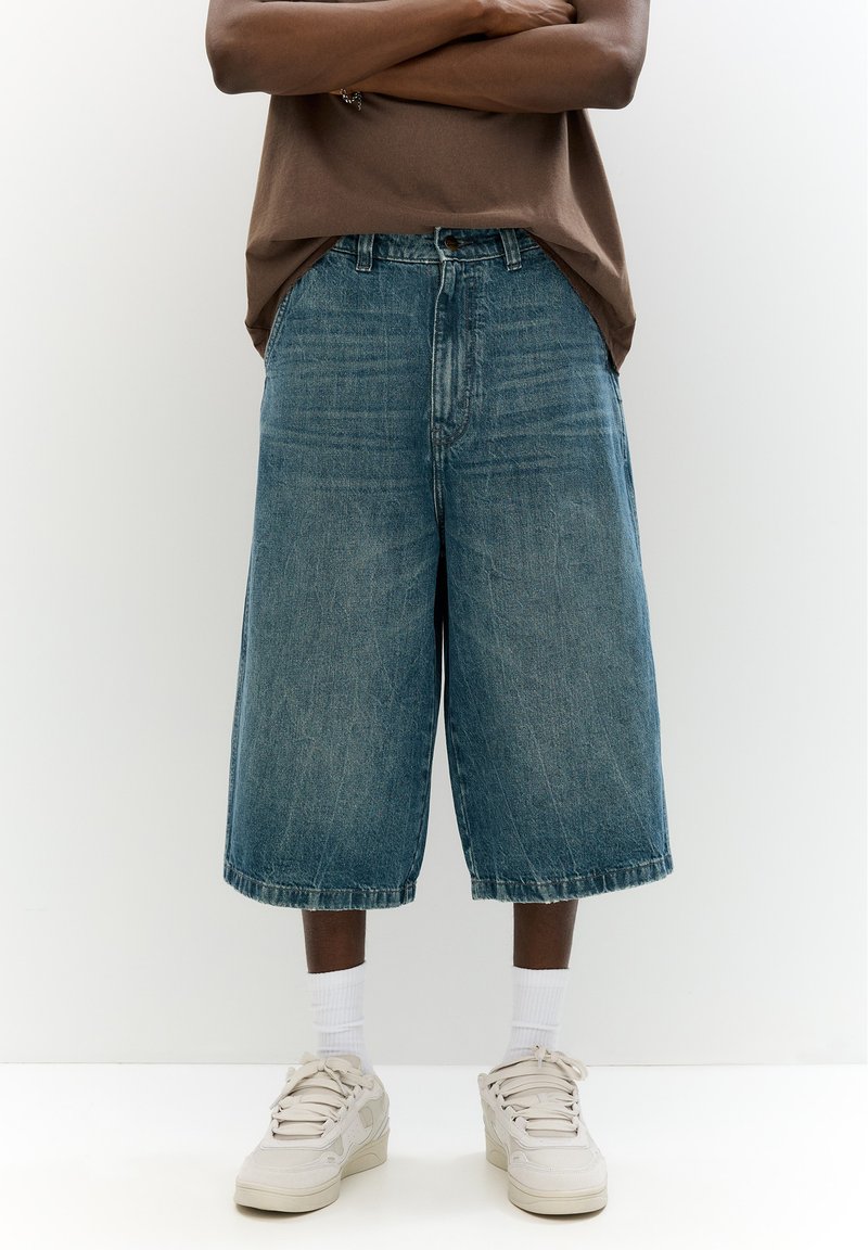 Shorts culotte in denim azzurro chiaro, caratterizzati da una vestibilità ampia, vita media e orlo sfilacciato, abbinati a calzini bianchi e sneakers beige.