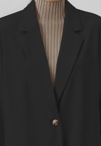 Blazer nero con un solo bottone d'oro, revers strutturati e taschino sul petto, sopra un dolcevita beige a coste. Tessuto liscio.