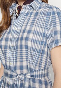 Modré a bílé kostkované šaty s krátkými rukávy, límečkem, knoflíkovou légou a pasem s opaskem pro nastavitelný střih.
