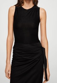 Schwarzer Bodysuit mit ärmellosem Design, aus glattem Stoff gefertigt. Kombiniert mit einem gerafften schwarzen Wickelrock mit einer Bindeschleife an der Seite.