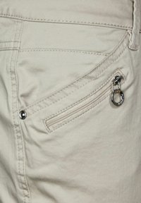 Close-up van een beige broekzak met zichtbare stiksels, een metalen klinknagel en een klein ritszakje met een ronde metalen trekker.