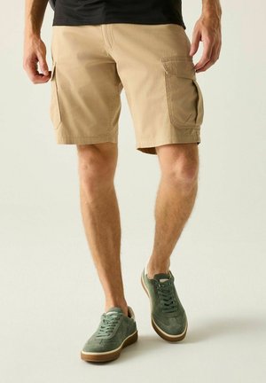 Uomo con pantaloncini cargo beige e sneakers verdi in camoscio, in piedi su sfondo bianco.