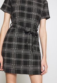 Svart och vit rutiga klänning med korta ärmar, midjeband, texturerad tyg och åtsittande siluett. Har kontrasterande rutmönster.