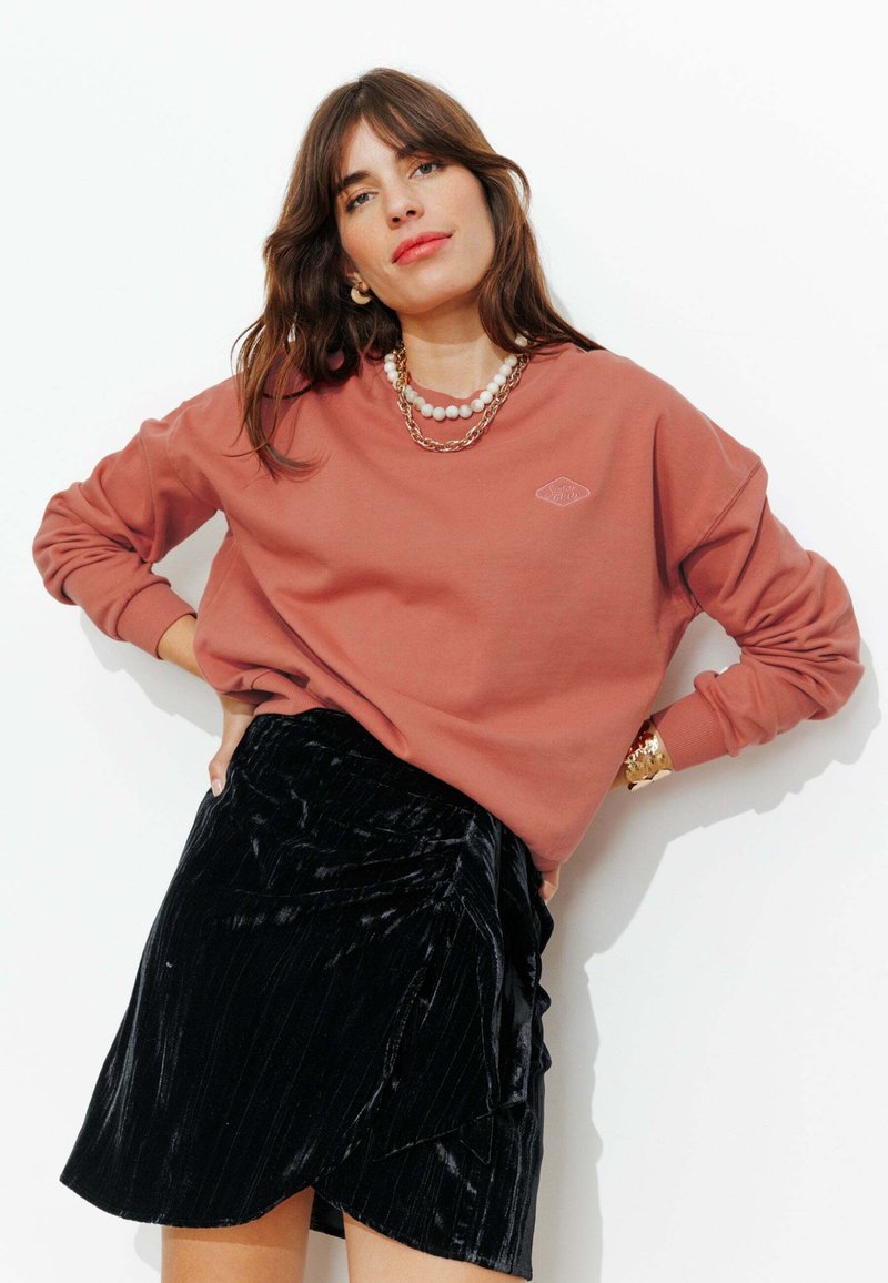 Sweat-shirt couleur rouille avec logo brodé, associé à une jupe portefeuille en velours noir présentant un design structuré et un côtelé subtil.