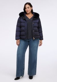 Navy-Puffermantel mit einer Kapuze aus Kunstfell, horizontal gestepptem Design und goldfarbenen Beschlägen, kombiniert mit einem grauen Oberteil und weit geschnittenen Jeans.