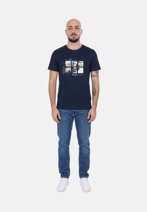 Uomo con tatuaggi che indossa una t-shirt grafica blu navy, jeans blu e sneakers bianche, in piedi di fronte su uno sfondo bianco semplice.
