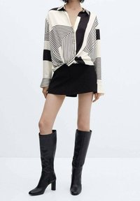 Chemise à manches longues à motif géométrique noir et blanc, associée à une jupe noire et des bottes en cuir noires montantes jusqu'aux genoux avec des talons carrés.