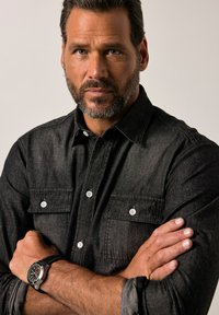 Camicia in denim nero con colletto a bottoni, due tasche sul petto e bottoni bianchi; abbinata a un orologio multifunzione con cinturino nero.
