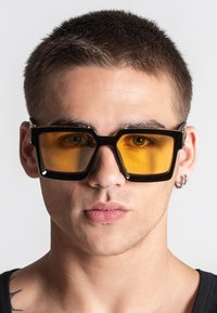 Occhiali da sole neri rettangolari con lenti gialle, montatura spessa e design geometrico. Presentano un accento argento sulla tempia.