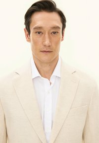 Beige linnen blazer met een notch-revers, enkele borstzak, gedragen over een wit overhemd met knopen. Glad oppervlak, getailleerd ontwerp, minimale details.