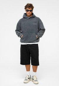 Grijze oversized hoodie met kangoeroezak, zwarte shorts en chunky witte sneakers. Model draagt donkere zonnebril en witte sokken.