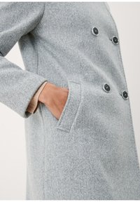 Cappotto in misto lana grigio con design doppiopetto, grandi bottoni neri e tasche laterali; presenta tessuto strutturato e vestibilità sartoriale.