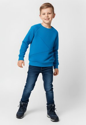 Młody chłopiec uśmiechający się, ubrany w niebieską bluzę, ciemne jeansy i czarne buty, stojący na tle jednolitego białego tła.