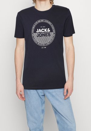 Młoda osoba ubrana w czarną koszulkę z logo Jack & Jones i jasnoniebieskie dżinsy, stojąca na jednolitym białym tle.