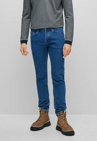 Man med blå jeans, bruna snörstövlar och en långärmad grå skjorta står mot en plain grå bakgrund.