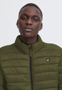 Gros plan d'une personne portant une veste matelassée zippée vert olive avec un petit logo sur le côté gauche de la poitrine et une boucle d'oreille créole à l'oreille gauche.