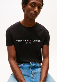 Ung mann iført en svart Tommy Hilfiger T-skjorte med logo og blå jeans mot en ensfarget hvit bakgrunn.